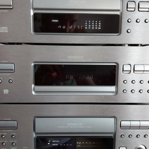Pu&ograve; includere: Un sistema stereo Kenwood argentato con un lettore CD, un sintonizzatore e un registratore a cassette. Il lettore CD ha un display digitale che mostra "no disc". Il sintonizzatore ha un display digitale che mostra "AM 12:00". Il registratore a cassette ha un display digitale che mostra "00:00".