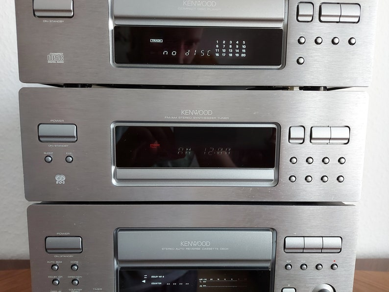 Pu&ograve; includere: Un sistema stereo Kenwood argentato con un lettore CD, un sintonizzatore e un registratore a cassette. Il lettore CD ha un display digitale che mostra "no disc". Il sintonizzatore ha un display digitale che mostra "AM 12:00". Il registratore a cassette ha un display digitale che mostra "00:00".
