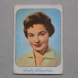 May include: A vintage trading card featuring a portrait of a woman with brown hair and blue eyes. The card has a blue background and the name "Mady Manstein" is written in blue ink at the bottom.