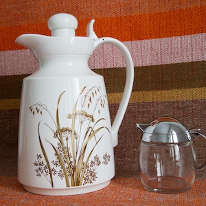 May include: A white ceramic thermal carafe with a floral design and a silver lid. The carafe has a brown and gold floral pattern with a design of grass and flowers. A clear glass creamer with a silver lid is also in the image.