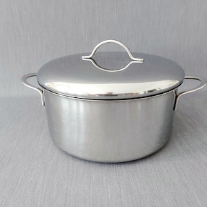 Puede incluir: Una olla de acero inoxidable pulido con tapa a juego. La olla cuenta con dos asas laterales curvas y una tapa redondeada con un asa central arqueada. La superficie del metal es reflectante y lisa, mostrando un acabado metálico limpio.