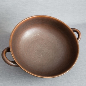 Könnte beinhalten: Eine braune Keramikschale mit zwei Schlaufengriffen. Die Schale hat eine runde Form und einen glatten Innenraum. Die Außenseite hat eine leicht strukturierte Oberfläche. Die Schale eignet sich zum Servieren von Speisen.