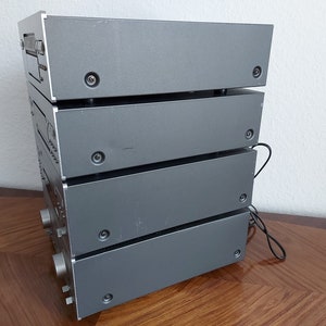 Pu&ograve; includere: Una pila di quattro componenti audio color argento, probabilmente un sistema stereo, con una superficie in legno in primo piano.