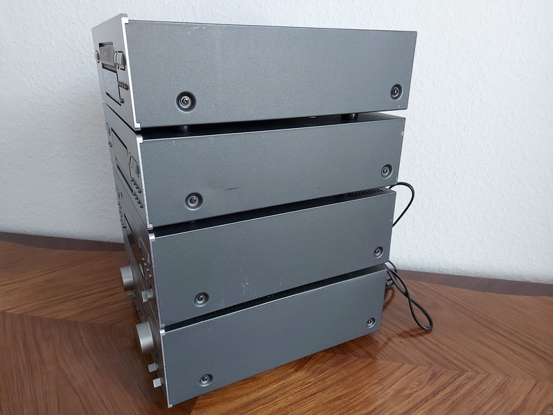 Pu&ograve; includere: Una pila di quattro componenti audio color argento, probabilmente un sistema stereo, con una superficie in legno in primo piano.