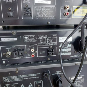 Pu&ograve; includere: Vista posteriore di un sistema stereo Kenwood nero con un lettore CD, un sintonizzatore e un amplificatore. Il sistema ha una piastra frontale argentata e un pannello di controllo nero. La parte posteriore del sistema ha vari jack di ingresso e uscita, tra cui antenna, altoparlanti e connessioni audio. Il sistema &egrave; etichettato "Kenwood Corporation" e "Made in Singapore".