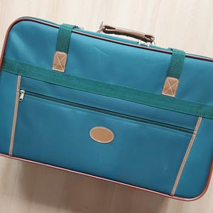 May include: Teal suitcase with brown trim and accents. The rectangular bag has a zipper pocket, a brown oval emblem, and green straps with brown leather-like patches. The handle is brown.