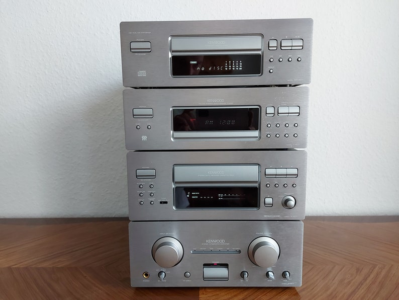Pu&ograve; includere: Un sistema stereo Kenwood argentato con un lettore CD, un registratore a cassette e un amplificatore. Il sistema ha un display digitale e diversi pulsanti.