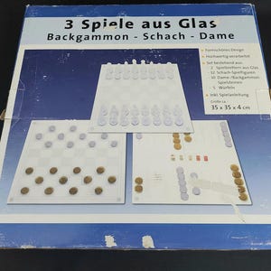 Könnte beinhalten: Eine Schachtel für ein Glasspielset mit Backgammon, Schach und Dame. Die Schachtel ist weiß mit blauen Akzenten und zeigt ein Bild der Spielbretter. Der Text auf der Schachtel lautet "3 Spiele aus Glas Backgammon - Schach - Dame".