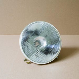 May include: A round, clear headlight with a white rim and a grid pattern on the glass. The headlight has a reflective interior and is mounted on a gold-colored bracket. The background is a plain, light brown surface.