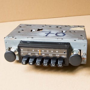 Può includere: Autoradio Ford vintage con manopole nere e frontalino nero. La radio ha un quadrante delle frequenze e diversi pulsanti. Il numero "70" è scritto sulla parte superiore della radio.