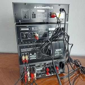 Pu&ograve; includere: Una pila di quattro componenti stereo neri con vari cavi e connettori sul retro. I componenti sono etichettati "Digital Output Optical", "Speaker Control", "FM Tuner", "CD Player", "ACC", "CAUTION" e "ATTENTION".