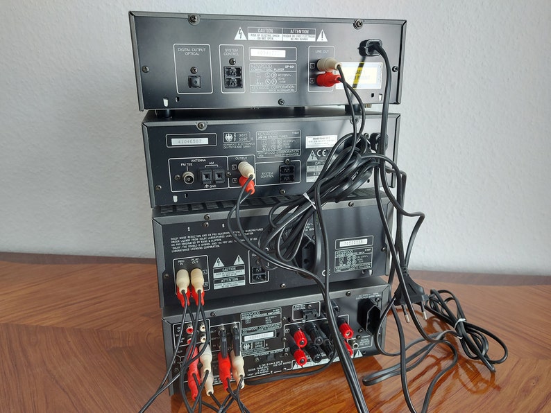 Pu&ograve; includere: Una pila di quattro componenti stereo neri con vari cavi e connettori sul retro. I componenti sono etichettati "Digital Output Optical", "Speaker Control", "FM Tuner", "CD Player", "ACC", "CAUTION" e "ATTENTION".