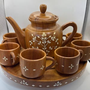 May include: A wooden tea set featuring a teapot with a lid, six matching cups, and a circular tray. The set is crafted from medium-brown wood and decorated with white inlaid floral and geometric patterns on the teapot, cups, and tray.