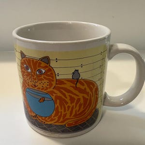 May include: A ceramic mug featuring a cartoon cat. The orange cat has a blue bowl and a small grey mouse. The mug has a white handle and rim, set against a yellow background with black lines.