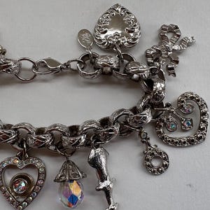 May include: Silver-toned charm bracelet with various decorative charms. Charms include hearts, bows, and a teardrop-shaped crystal. The bracelet has a textured chain link design and a lobster clasp closure.