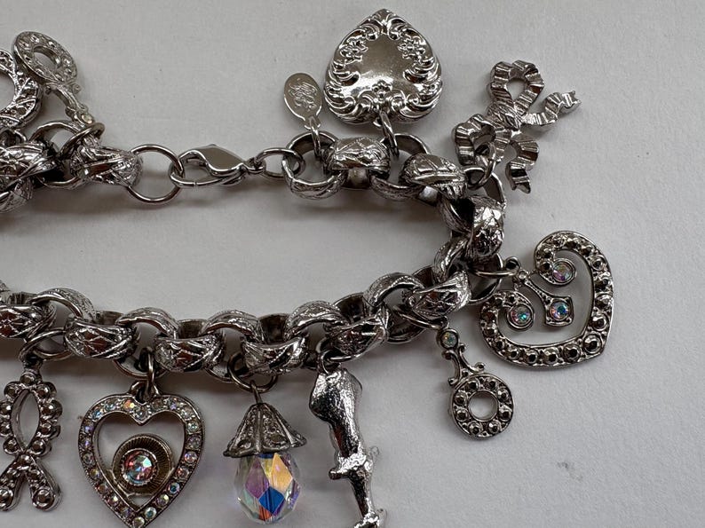 May include: Silver-toned charm bracelet with various decorative charms. Charms include hearts, bows, and a teardrop-shaped crystal. The bracelet has a textured chain link design and a lobster clasp closure.