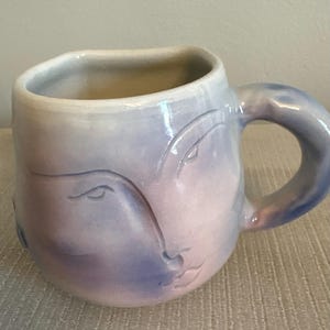 Könnte beinhalten: Eine Keramiktasse mit einem Gesichtsdesign. Die Tasse ist überwiegend hellblau und rosa, mit einem gebogenen Henkel. Das Gesicht ist dezent in die Keramik gemeißelt, mit zarten Linien, die die Merkmale definieren. Die Tasse hat ein einzigartiges, künstlerisches Aussehen.