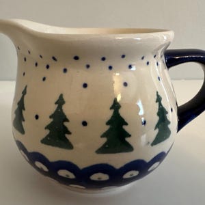 May include: A ceramic creamer with a white base, featuring a dark blue handle and a spout. The creamer is decorated with green Christmas trees and blue polka dots. A dark blue scalloped pattern encircles the base.
