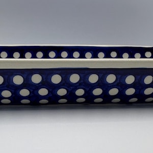 May include: A rectangular ceramic baking dish featuring a deep blue glaze decorated with a repeating pattern of small white polka dots. The dish has a thick, cream-coloured rim that extends around the top edge, providing a contrast to the blue.