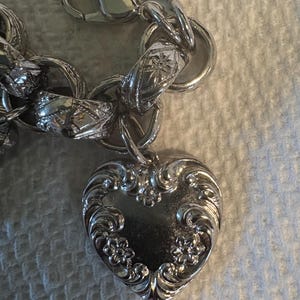May include: A silver-toned charm bracelet with a heart-shaped pendant. The heart pendant is intricately detailed with floral and scroll designs. The bracelet features a lobster clasp and other decorative links.