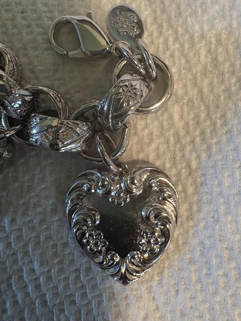 May include: A silver-toned charm bracelet with a heart-shaped pendant. The heart pendant is intricately detailed with floral and scroll designs. The bracelet features a lobster clasp and other decorative links.