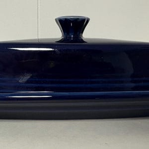 Op de afbeelding: Een rechthoekige botervloot van diep marineblauw keramiek met een bijpassend deksel. Het deksel heeft een centrale knop als handvat en subtiele horizontale reliëflijnen langs de basis. De schaal heeft een brede, platte rand en een gladde afwerking.