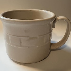 May include: A beige ceramic mug with a rounded handle. The mug features a textured, brick-like pattern around its body. The interior and handle match the exterior color. The mug is designed for hot beverages.