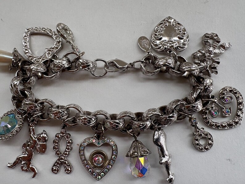 May include: Silver charm bracelet with various charms, including a heart, bow, and cherub. The bracelet features a chunky chain and a mix of textures and embellishments. Some charms have colorful accents.
