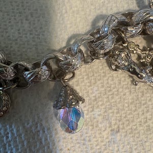 May include: Silver-toned charm bracelet with a large link chain. A teardrop-shaped charm with iridescent facets hangs from the chain. Other charms are visible, including a round charm with a floral design.