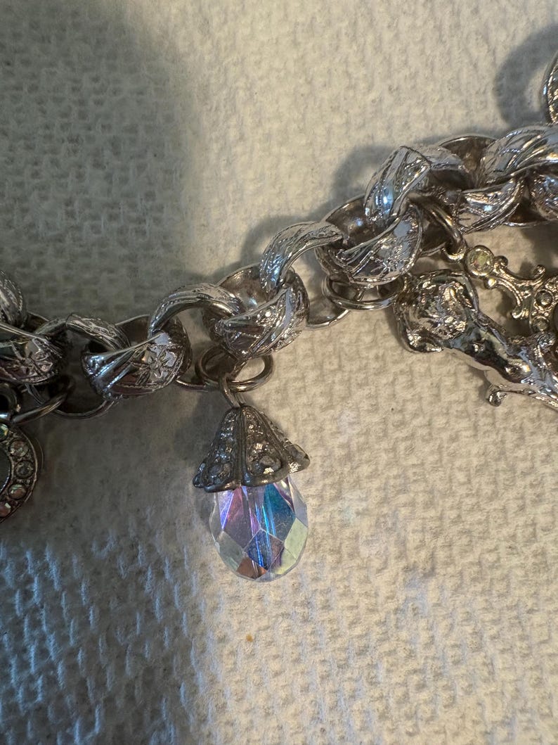 May include: Silver-toned charm bracelet with a large link chain. A teardrop-shaped charm with iridescent facets hangs from the chain. Other charms are visible, including a round charm with a floral design.