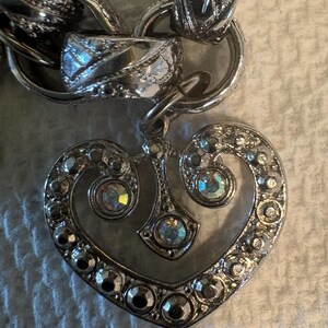 May include: Silver-toned heart-shaped pendant with clear crystal accents, attached to a chunky chain. The heart pendant features a detailed design with multiple crystals around the edges and in the center, creating a sparkling effect.