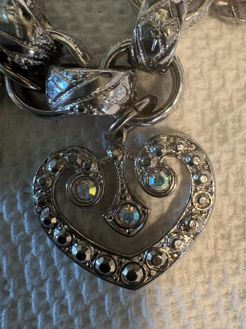 May include: Silver-toned heart-shaped pendant with clear crystal accents, attached to a chunky chain. The heart pendant features a detailed design with multiple crystals around the edges and in the center, creating a sparkling effect.