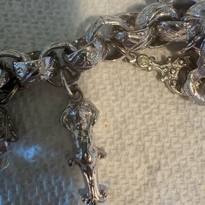 May include: Close-up of a silver-toned charm bracelet with ornate links and decorative charms. The charms include a detailed figure and other decorative elements. The bracelet is set against a textured, off-white background.