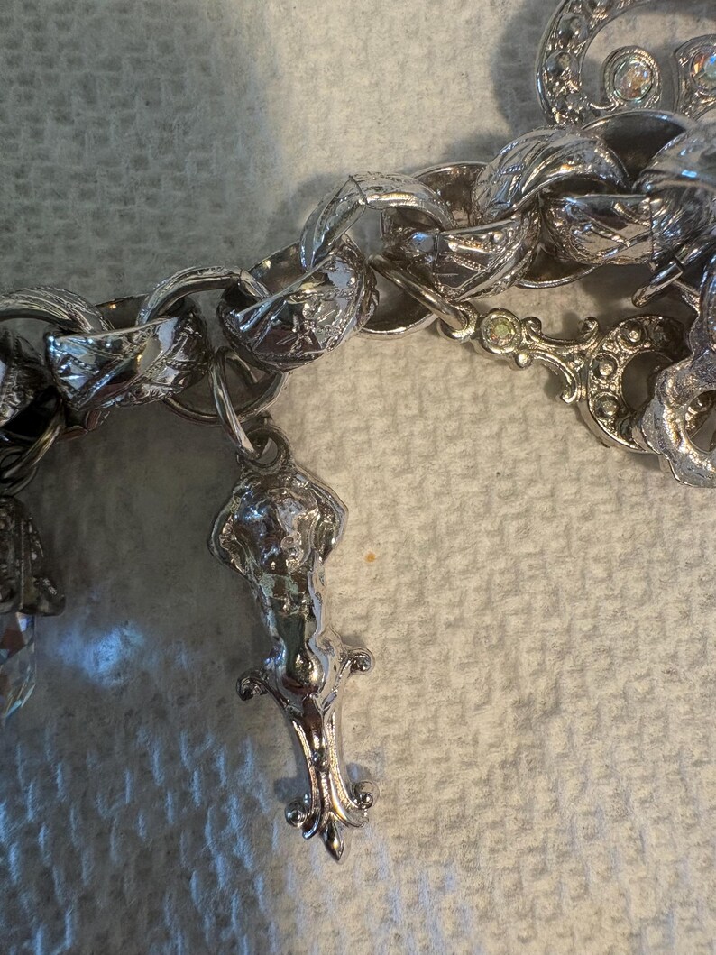 May include: Close-up of a silver-toned charm bracelet with ornate links and decorative charms. The charms include a detailed figure and other decorative elements. The bracelet is set against a textured, off-white background.