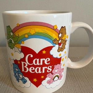 Pode incluir: Caneca de cerâmica branca com personagens dos Ursinhos Carinhosos, um arco-íris e estrelas. A caneca tem um grande coração vermelho com as palavras "Care Bears" em branco. A alça é branca.