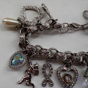 May include: Silver-toned charm bracelet with various charms, including a pearlescent bead, a heart-shaped charm, a cherub, and a heart-shaped charm with iridescent rhinestones. The bracelet has a textured chain link design.
