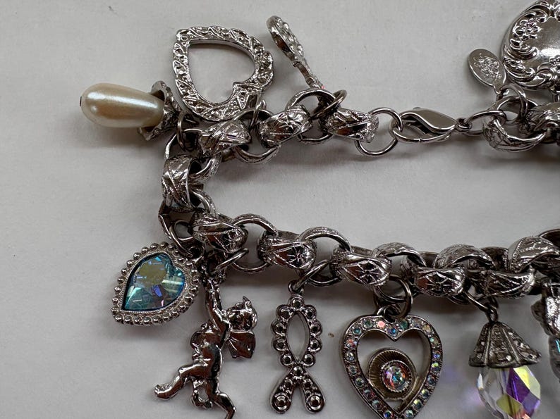 May include: Silver-toned charm bracelet with various charms, including a pearlescent bead, a heart-shaped charm, a cherub, and a heart-shaped charm with iridescent rhinestones. The bracelet has a textured chain link design.