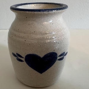 Puede incluir: Una vasija de gres con esmalte blanquecino moteado y un motivo de corazón pintado en azul oscuro en la parte frontal. El recipiente tiene un cuerpo ancho y redondeado, un borde ligeramente acampanado con acabado azul oscuro y textura rústica.