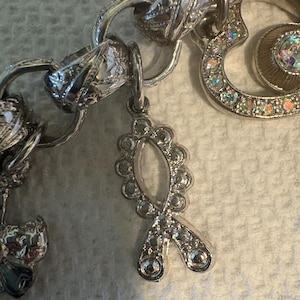 May include: Close-up of a silver charm bracelet with various charms. The bracelet features a ribbon charm adorned with clear rhinestones. Other charms include a heart-shaped charm with iridescent stones and a decorative link.