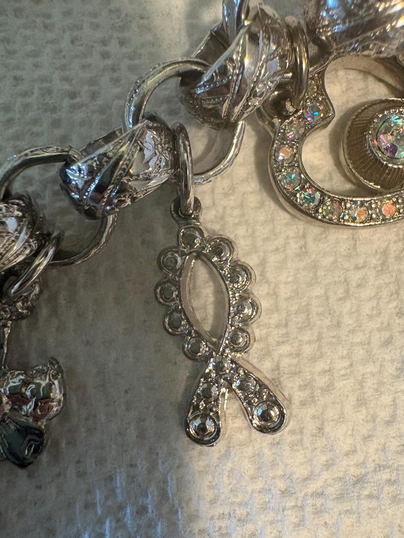 May include: Close-up of a silver charm bracelet with various charms. The bracelet features a ribbon charm adorned with clear rhinestones. Other charms include a heart-shaped charm with iridescent stones and a decorative link.