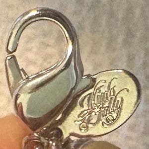 May include: Close-up of a silver-toned jewelry clasp with a small, heart-shaped tag. The tag features an ornate, stylized inscription. The clasp is open, revealing the connecting mechanism.
