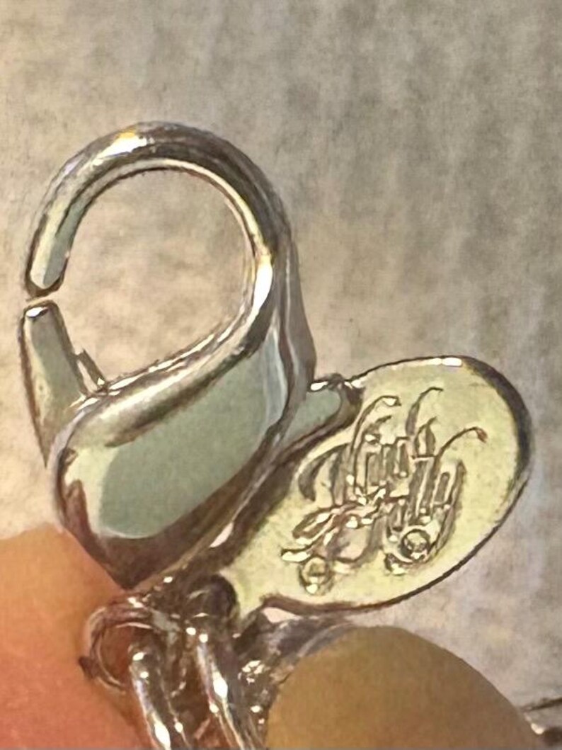 May include: Close-up of a silver-toned jewelry clasp with a small, heart-shaped tag. The tag features an ornate, stylized inscription. The clasp is open, revealing the connecting mechanism.