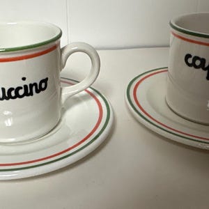 May include: Two white ceramic cups and matching saucers, each featuring the word cappuccino in black cursive script. The rims of the cups and the edges of the saucers are decorated with thin, concentric red and green circular bands.