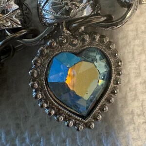 May include: Close-up of a silver-toned bracelet with a heart-shaped pendant. The pendant features a faceted, iridescent stone in a heart shape, surrounded by a beaded silver frame. The bracelet links have ornate detailing.