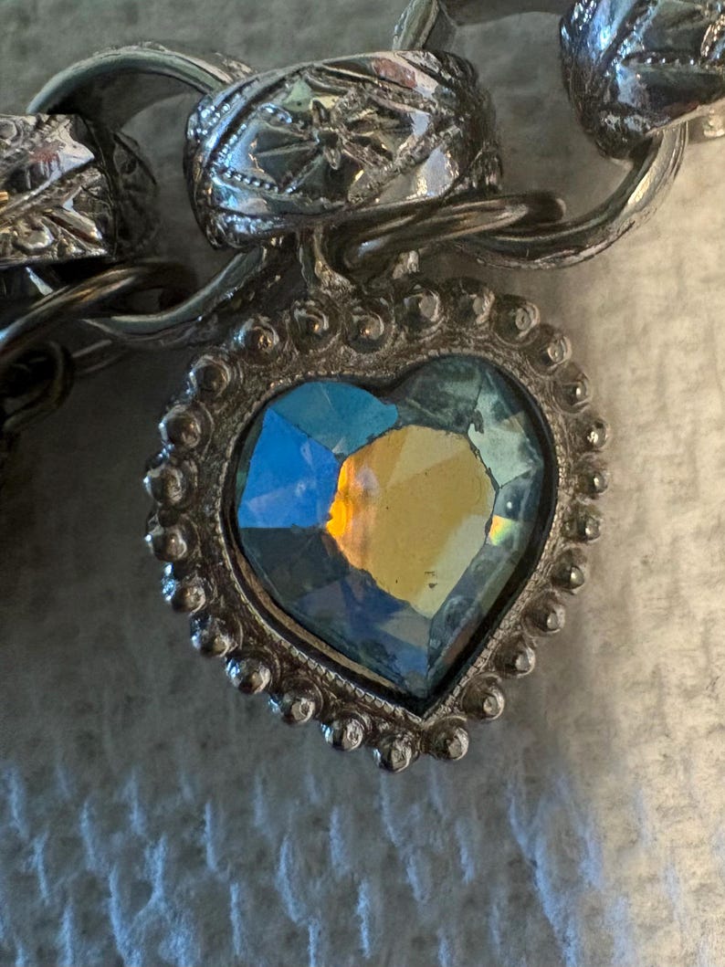 May include: Close-up of a silver-toned bracelet with a heart-shaped pendant. The pendant features a faceted, iridescent stone in a heart shape, surrounded by a beaded silver frame. The bracelet links have ornate detailing.