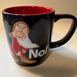 May include: Black ceramic mug with a red interior, featuring an illustration of Grumpy from Snow White. The character is shown in his red and brown outfit with a grumpy expression. The word "No!" is printed on the side of the mug.
