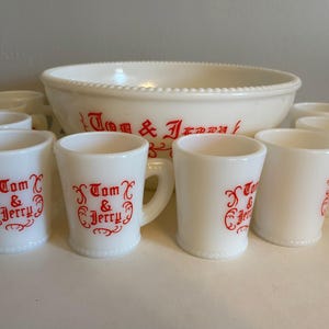 May include: A white milk glass punch bowl and matching mugs set featuring a beaded rim design. Each piece is decorated with the red text Tom & Jerry in a stylised font. The set includes one large bowl and several small handled mugs.