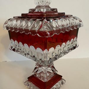 May include: A clear and ruby red glass candy dish with a lid. The dish has a square shape with scalloped edges and a pedestal base. The lid is topped with a decorative finial. The glass is cut with geometric patterns.