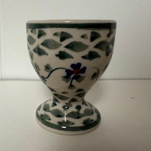 May include: A ceramic egg cup featuring a cream-colored base with a repeating pattern of dark green leaf-like shapes. The design includes a central blue flower with a red center, accented by thin blue stems and small gray leaves around the middle.