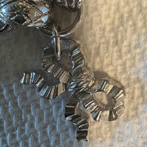 May include: A close-up of a silver-colored charm in the shape of a bow. The bow is attached to a silver chain. The charm has a textured surface with detailed ridges and curves, reflecting light.